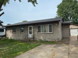 A single-story, horizontal-sided brown house with a white door, large front window, and an attached garage to the right.