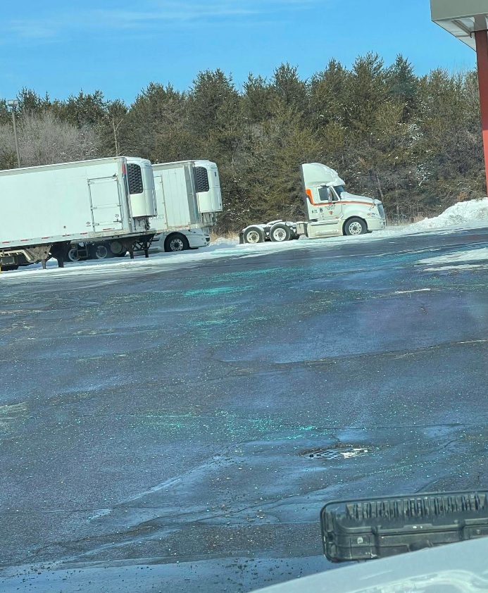 Two semi-trailers parked in a snowy lot with a white tractor unit driving past them near a line of trees.