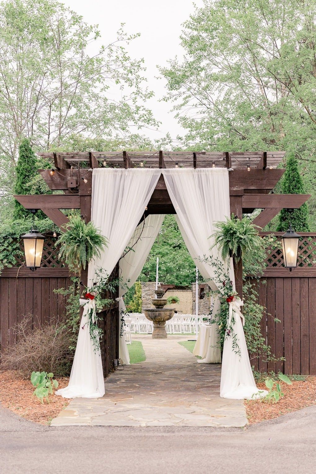 A wooden archway with white curtains and ferns is decorated for a wedding ceremony.