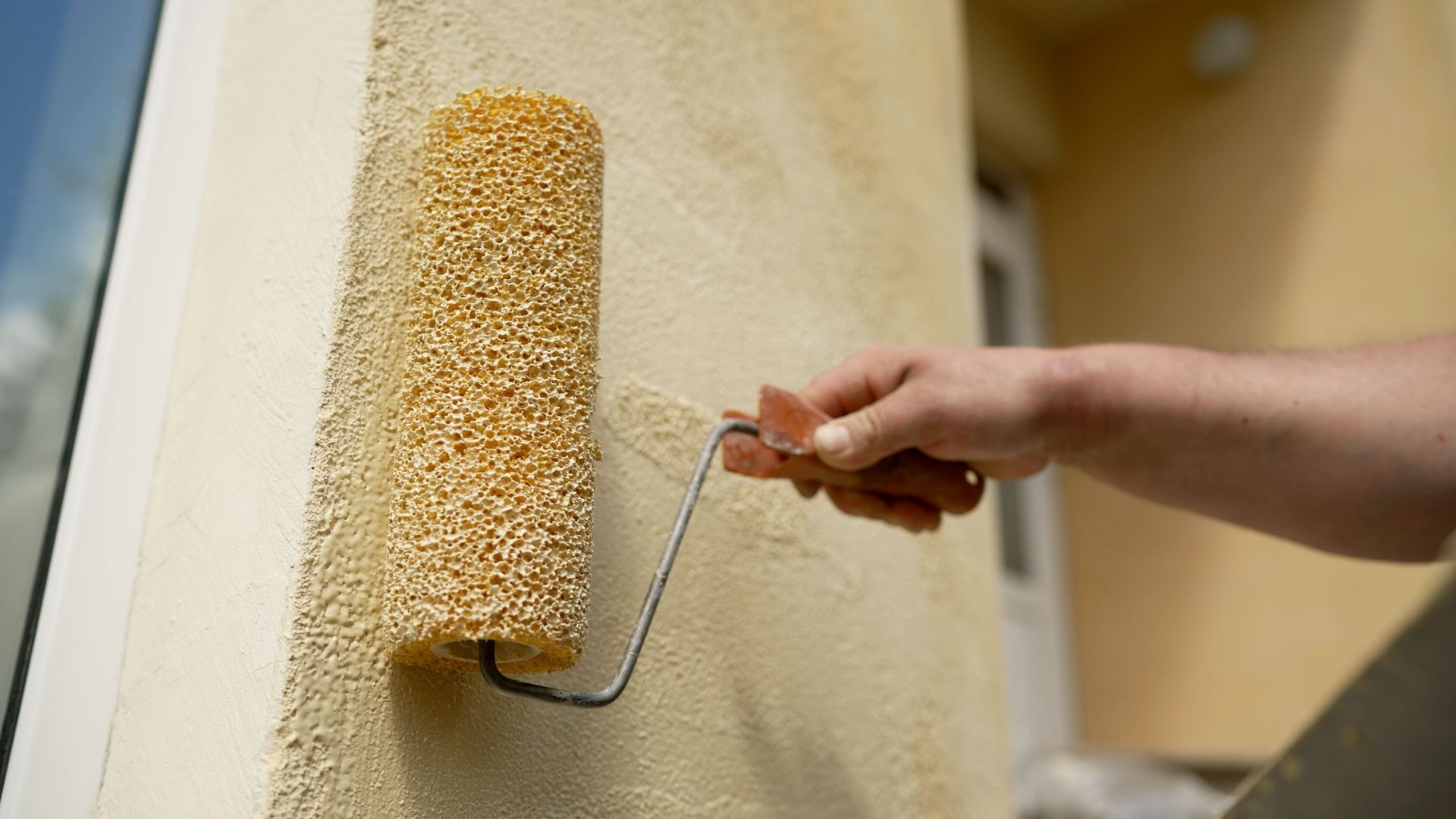 Person rolling textured coating onto a stucco wall; tan roller and wall.