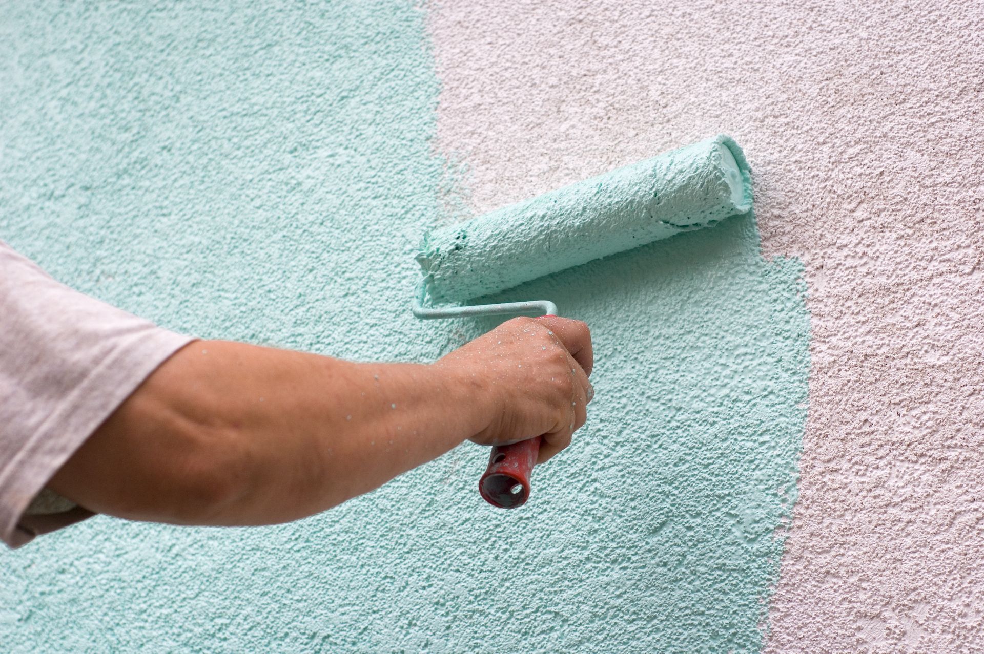 Person painting a stucco wall with a teal paint roller.