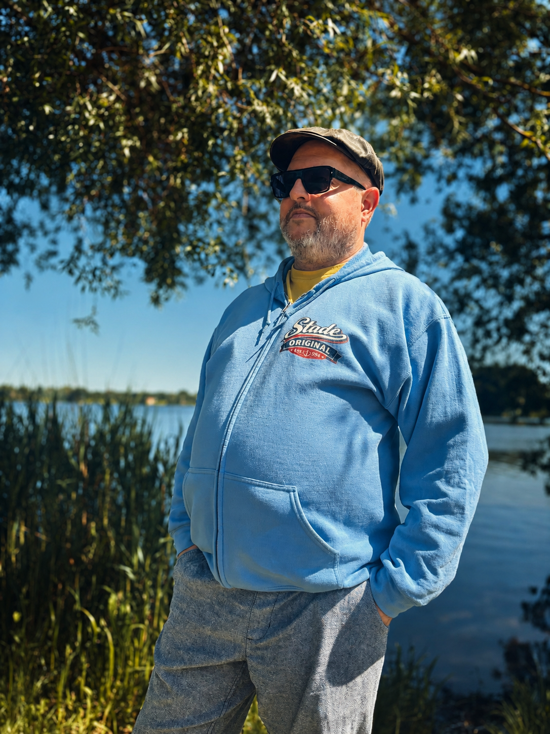 Ein Mann in orangefarbenem Kapuzenpulli steht unter klarem, blauem Himmel auf sandigen Dünen und blickt zur Seite.