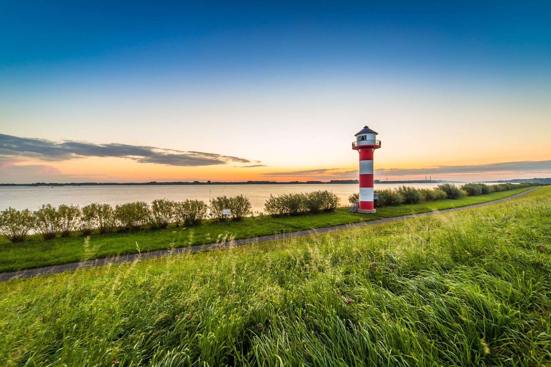 Ein rot-weißer Leuchtturm neben einem Fluss bei Sonnenuntergang, im Vordergrund grünes Gras und ruhiges Wasser