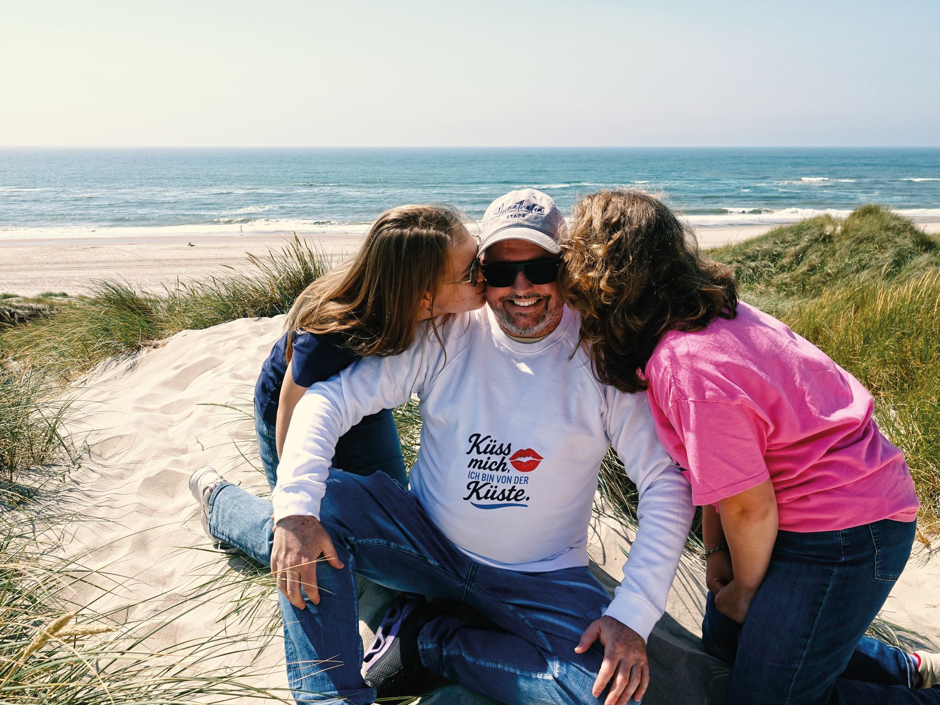 Drei Menschen sitzen lächelnd an einem Sandstrand, im Hintergrund die Wellen des Ozeans und Dünen.