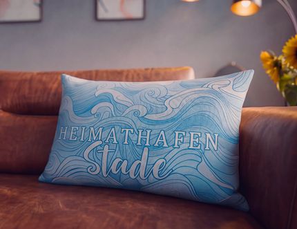 Blaues Dekokissen auf einem braunen Sofa mit der Aufschrift „Heimat Hafen Stadt“, im Hintergrund eine verschwommene Lampe und Blumen.
