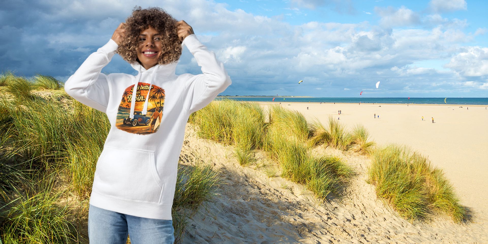 Eine Person in einem weißen Sweatshirt steht an einem Sandstrand mit Dünen und blauem Wasser, die Hände im Haar, unter einem sonnigen Himmel