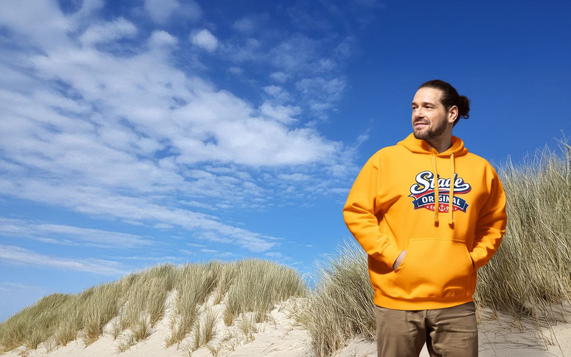 Eine Person in einem orangefarbenen Kapuzenpullover steht auf sandigen Dünen unter einem blauen Himmel mit Wolken.