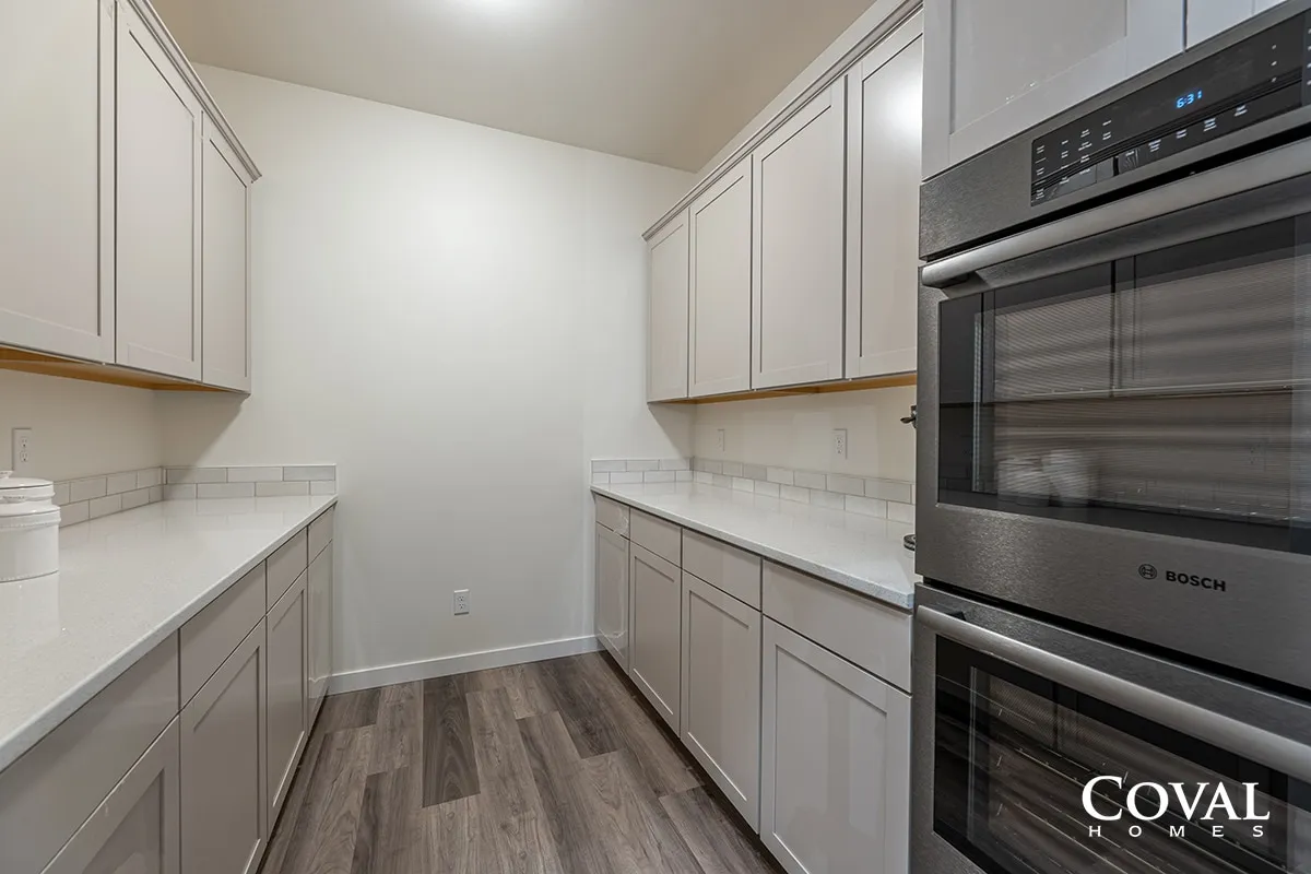 Bright galley kitchen with white cabinets, marble counters, and built-in stainless steel ovens.