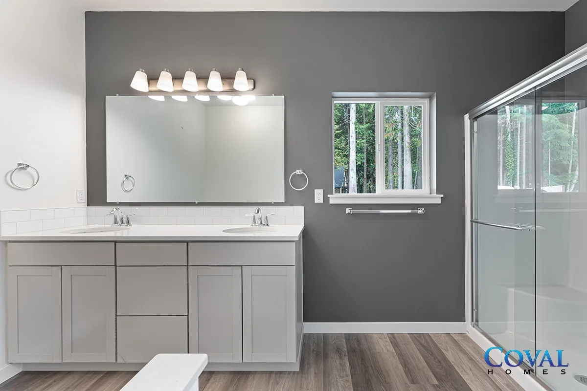 Modern bathroom with gray walls, double vanity, large mirror, window, and glass shower enclosure