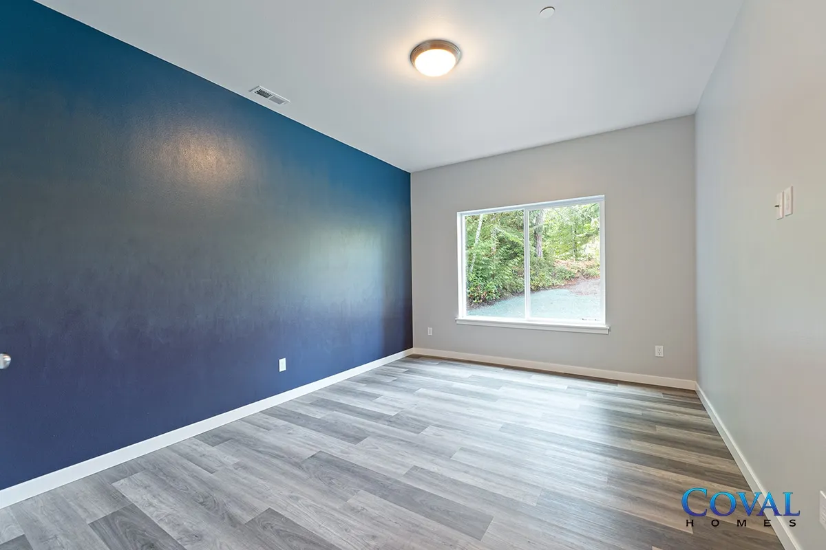 Empty bedroom with blue accent wall, gray flooring, and a window overlooking greenery.
