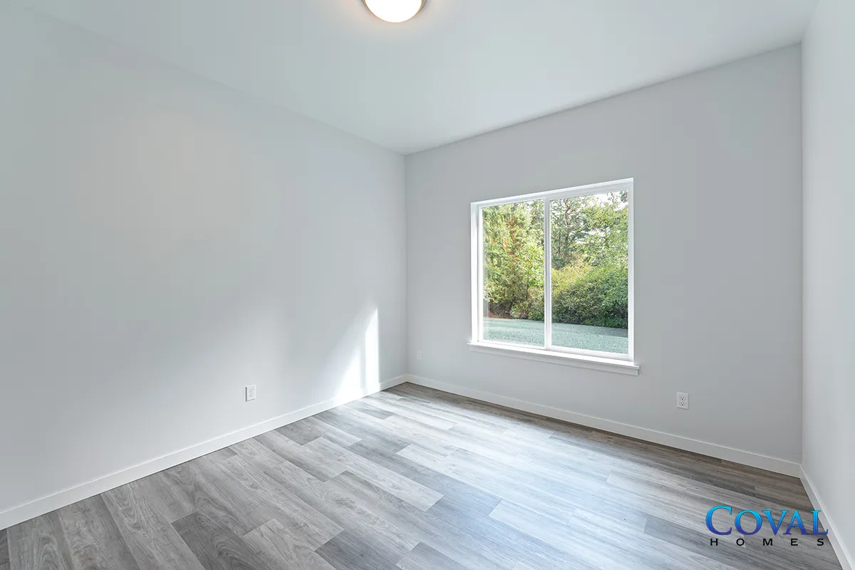 Empty bright bedroom with white walls, gray wood-look floor, and a window showing greenery