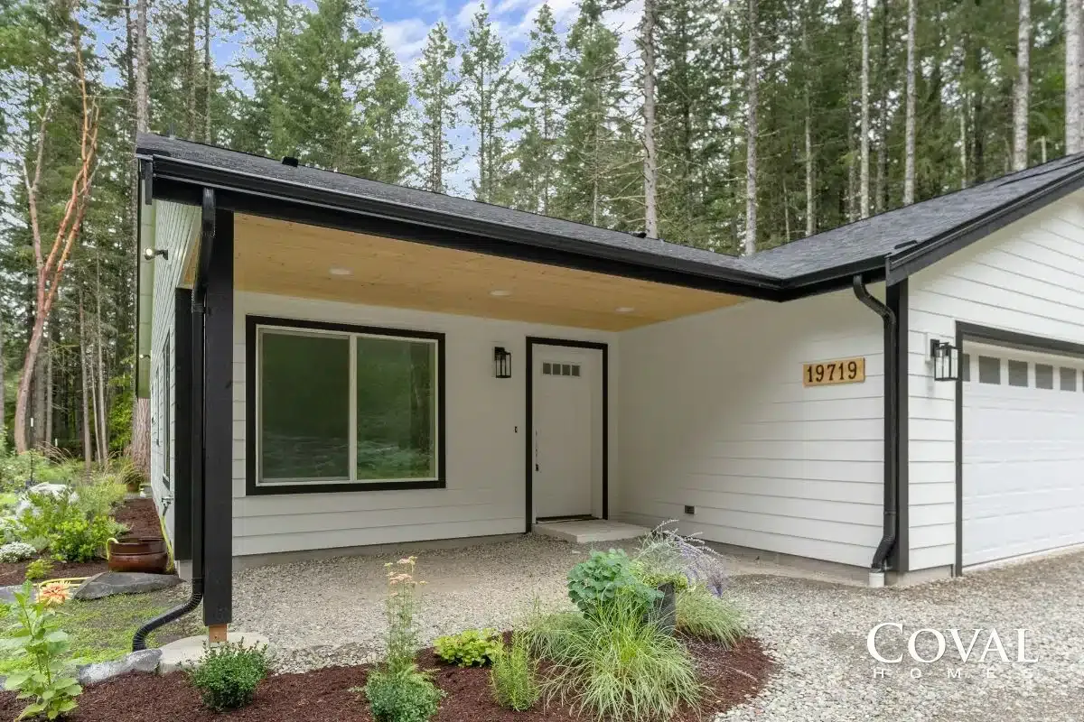 Modern house exterior with covered entry, white siding, black trim, and forest backdrop