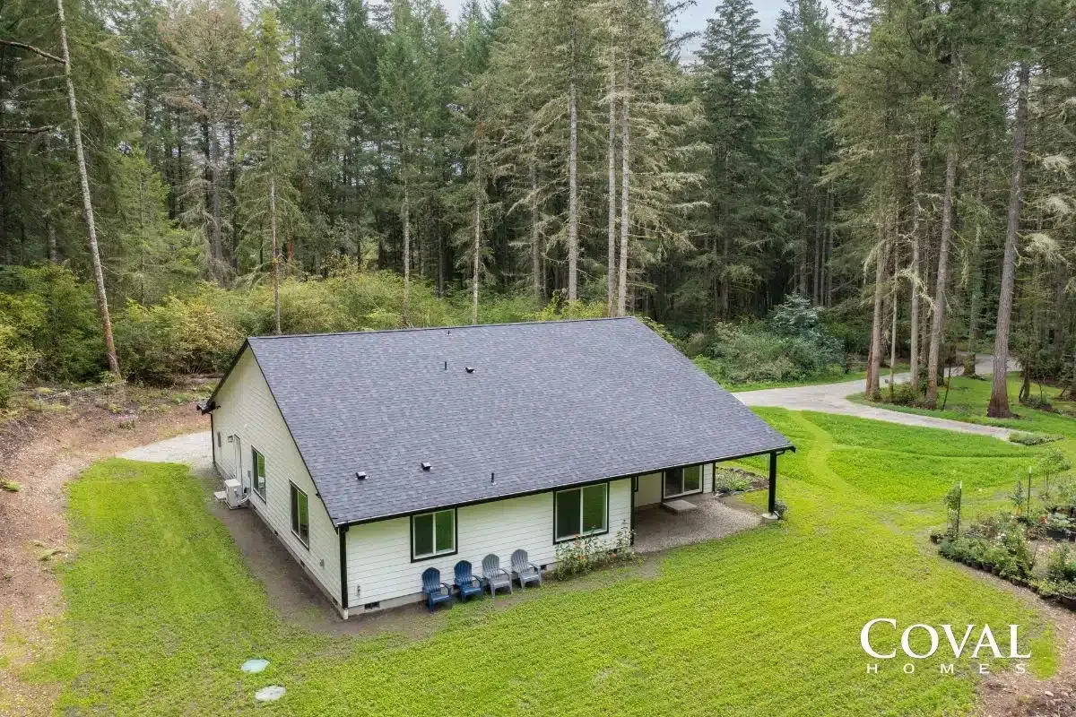 Aerial view of a small white house with gray roof, green lawn, and forest surrounding it