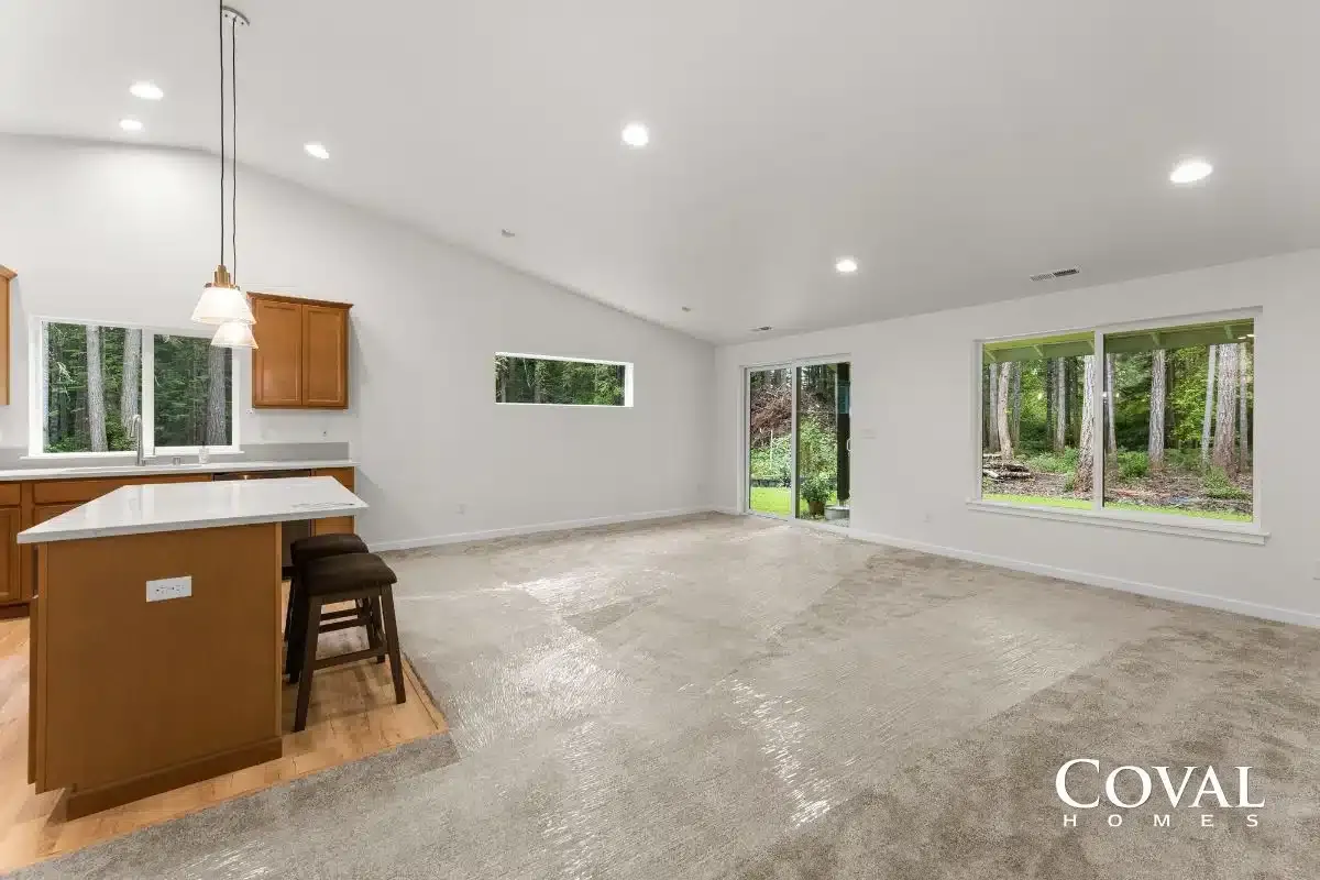 Bright open living room with kitchen island, carpet, recessed lights, and large windows overlooking trees