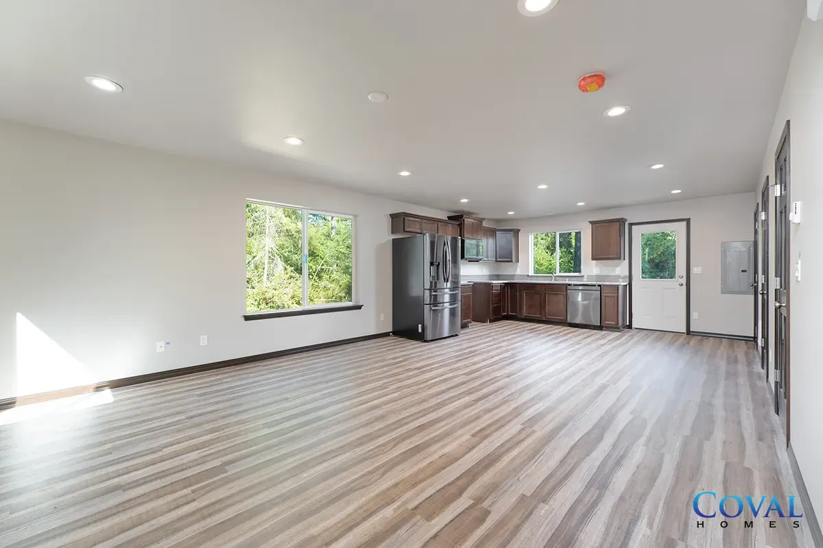 Bright open empty living area with wood-look floors, white walls, large windows, and kitchen in back