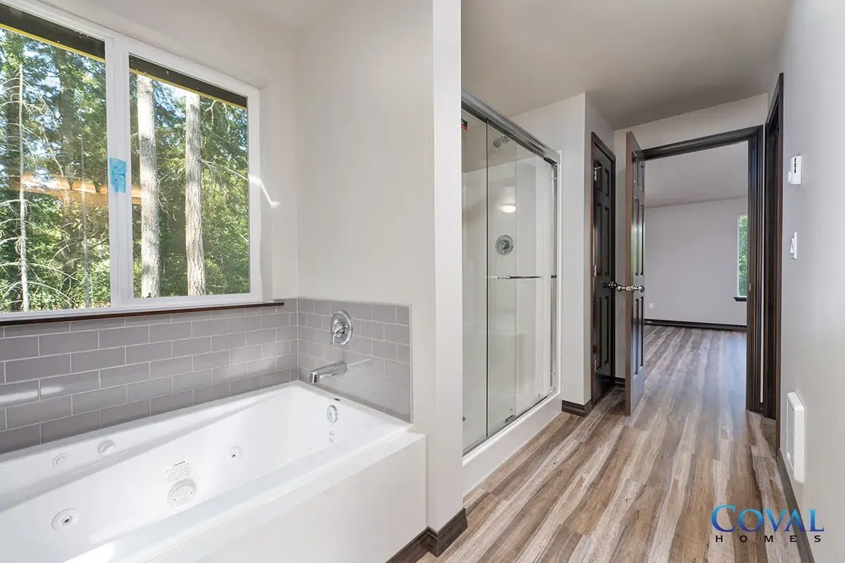 Bright bathroom with a white soaking tub, window, and gray tile leading to a hallway with wood floors