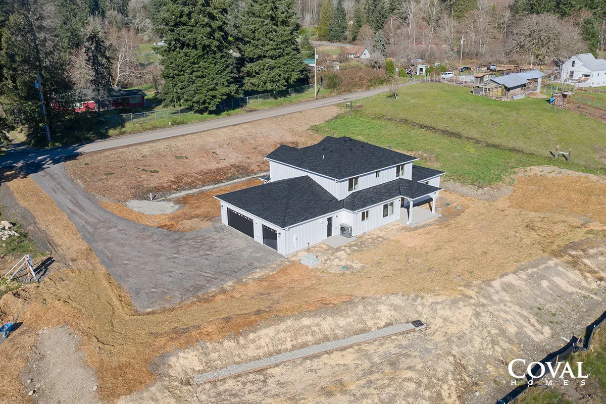 Aerial view of a newly built gray house with dark roof on a cleared lot beside woods and a driveway.