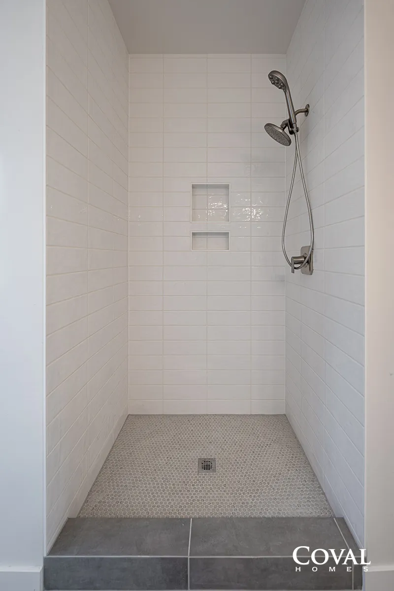 White tiled shower stall with built-in shelf, rainfall showerhead, and mosaic floor tiles.