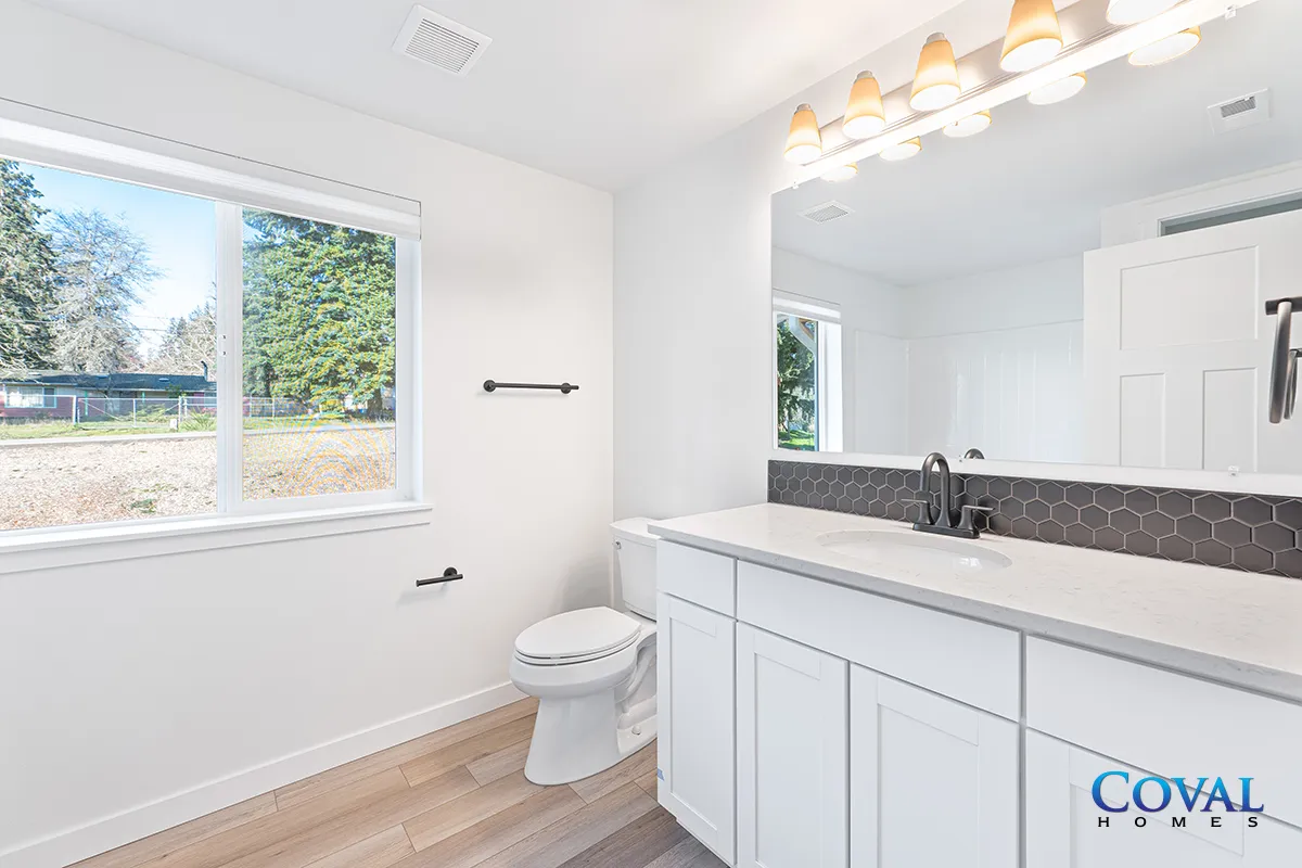 Bright bathroom with white vanity, large mirror, toilet, window, and light wood flooring