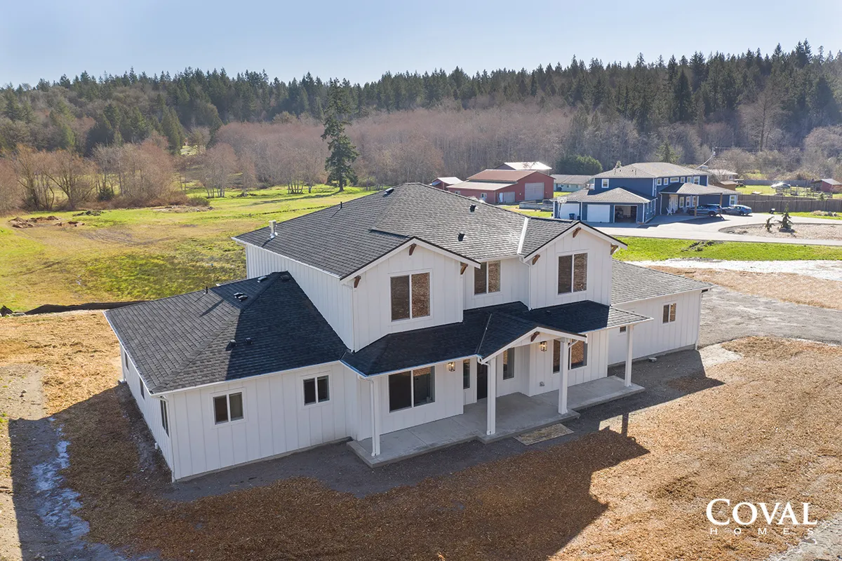 Aerial view of a large white house with dark roof on a cleared lot near woods and neighboring homes.