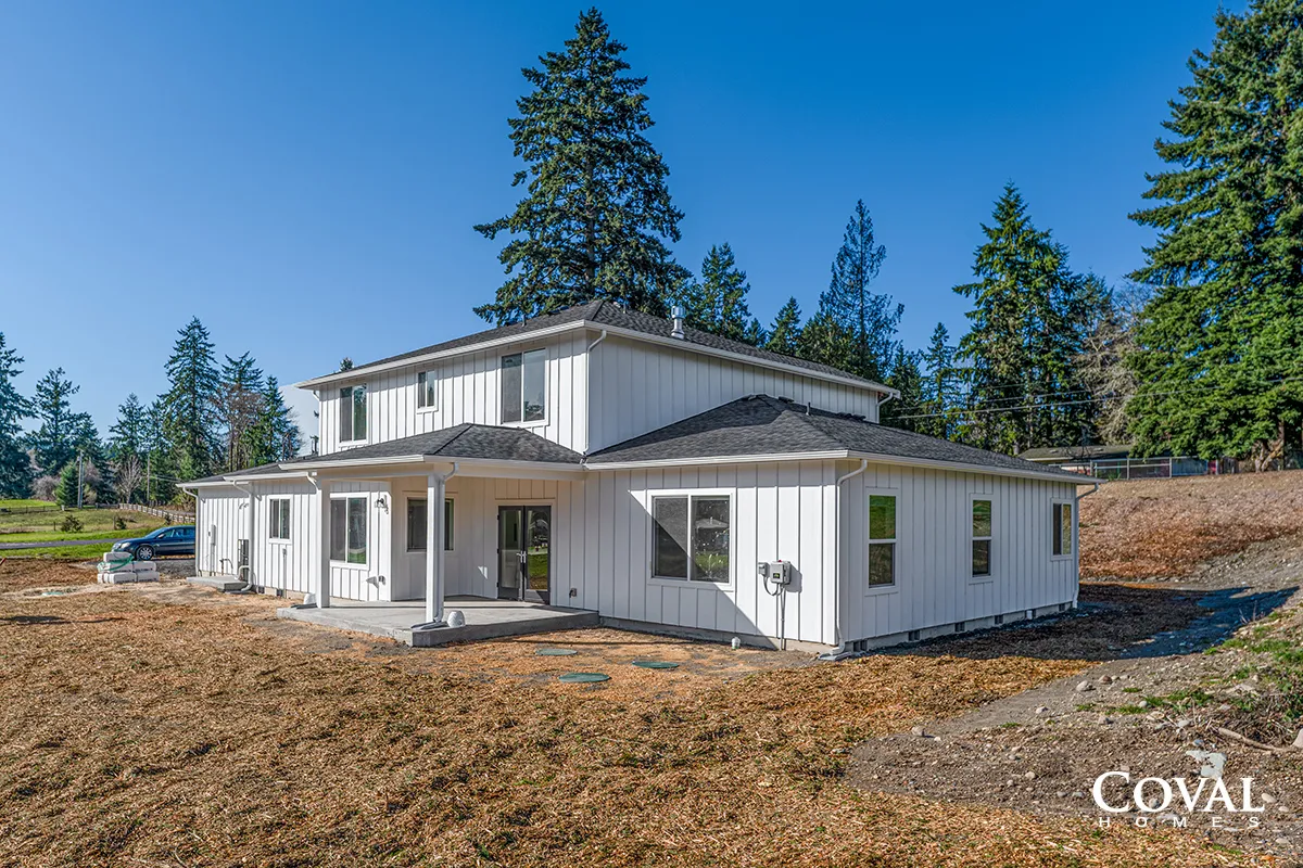 White two-story house with a covered porch on a barren lot, surrounded by tall evergreen trees under a blue sky