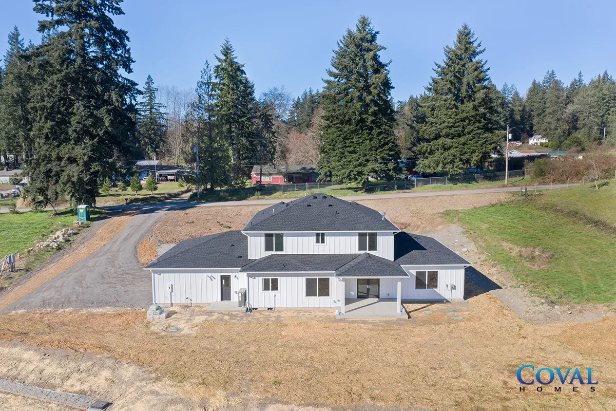 Aerial view of a gray house with a dark roof on a grassy hillside, surrounded by trees and open land