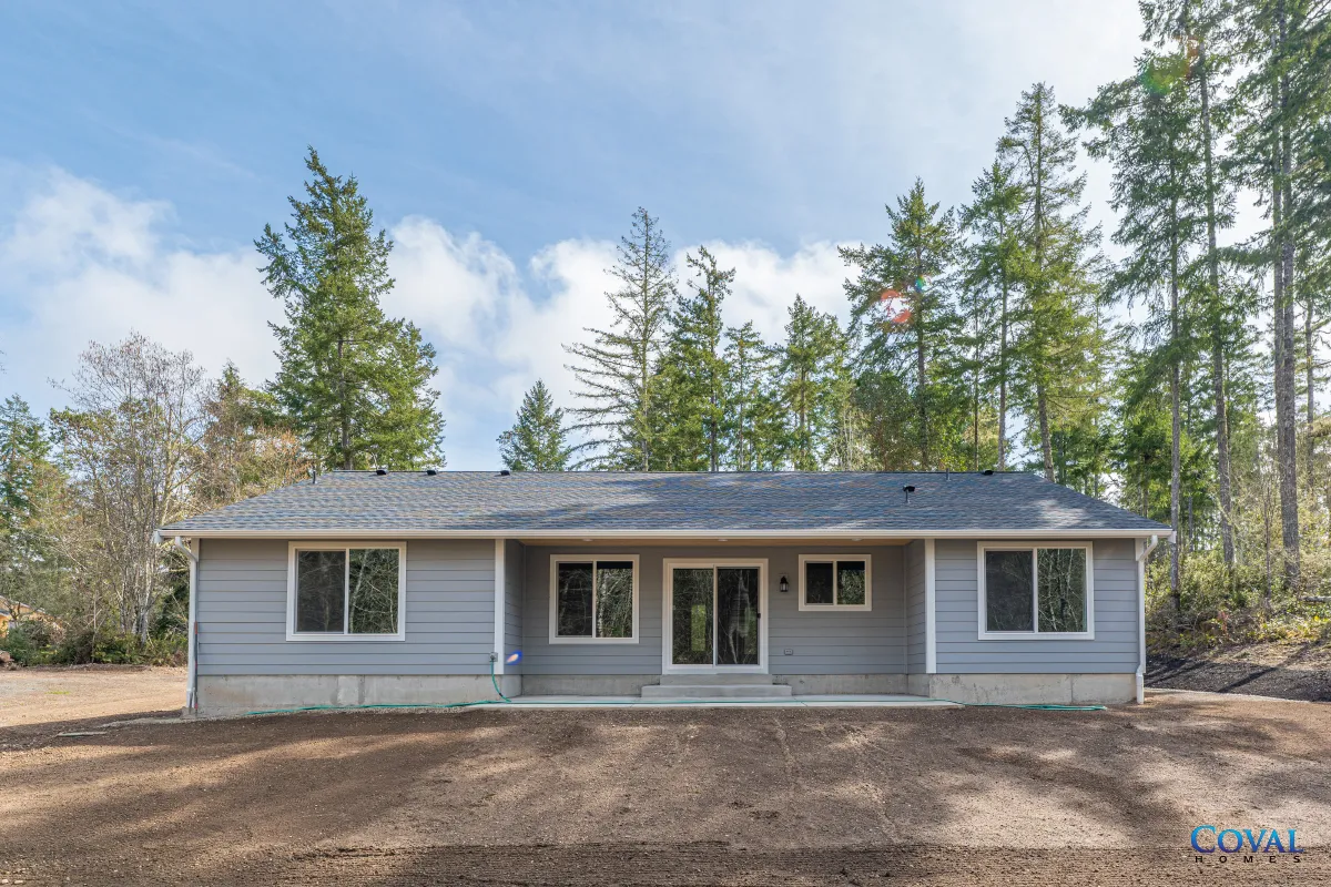 Single-story gray house with front porch, surrounded by trees on a bare lot under a partly cloudy sky