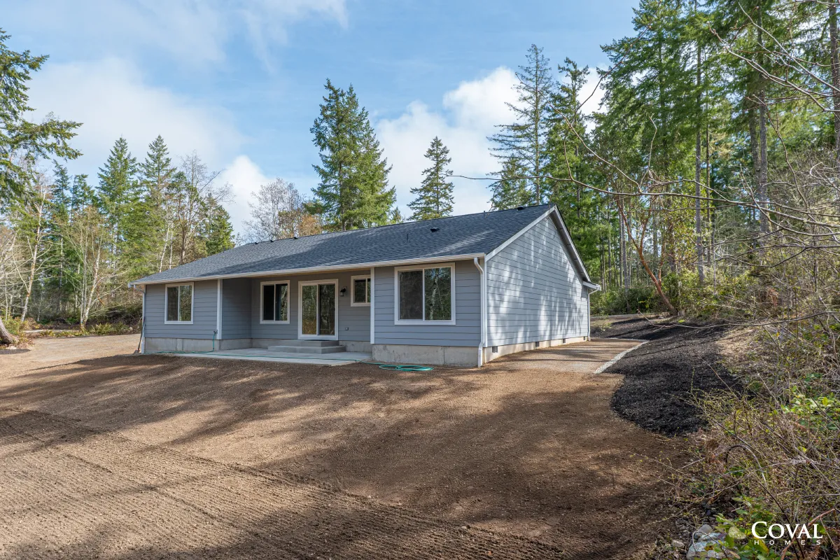 Gray single-story house with white trim, set among tall trees on a wooded lot under a blue sky