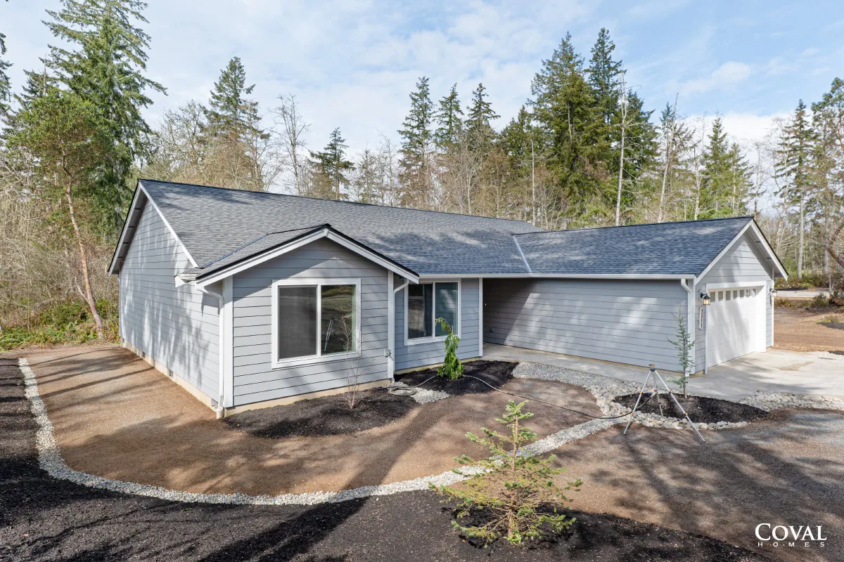 Gray single-story house with attached garage, dark roof, and wooded yard on a sunny day