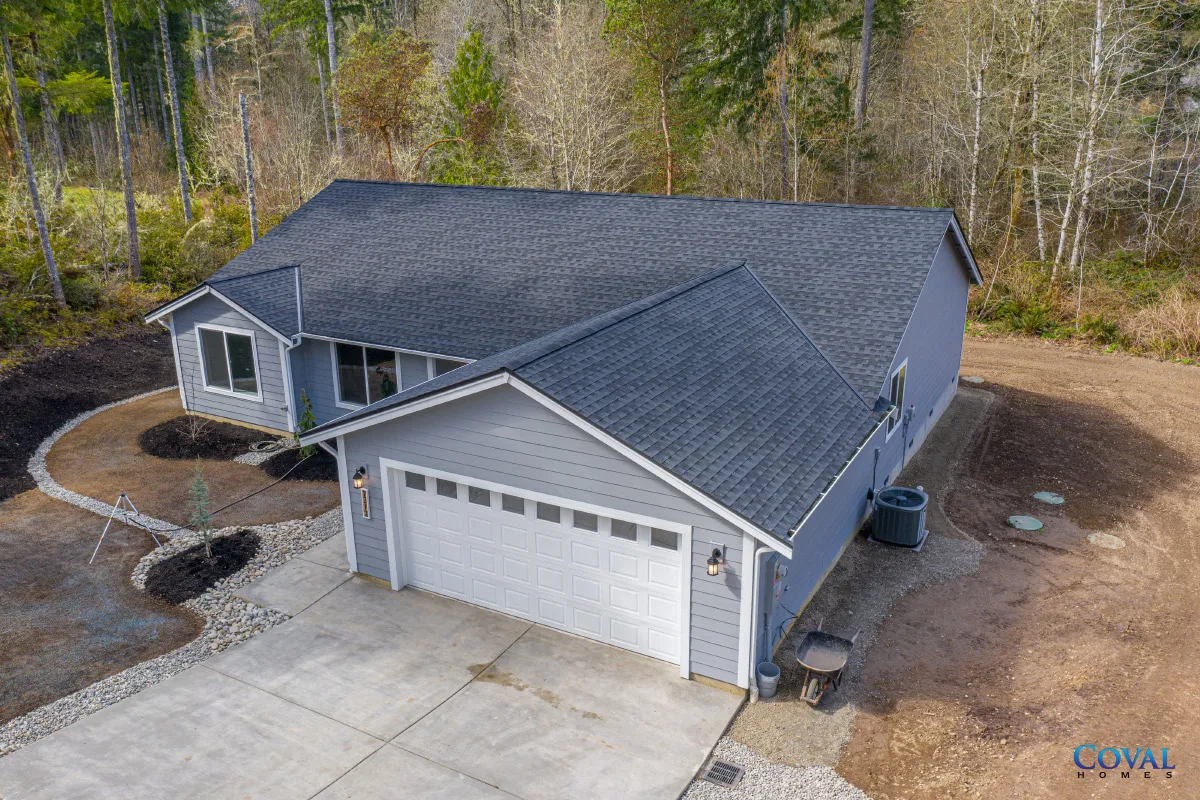 Aerial view of a gray suburban house with a two-car garage and driveway, surrounded by trees.