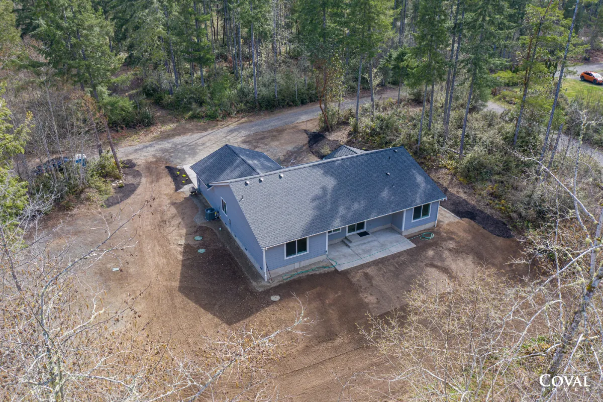 Aerial view of a gray house with a dark roof beside a wooded yard and dirt driveway.