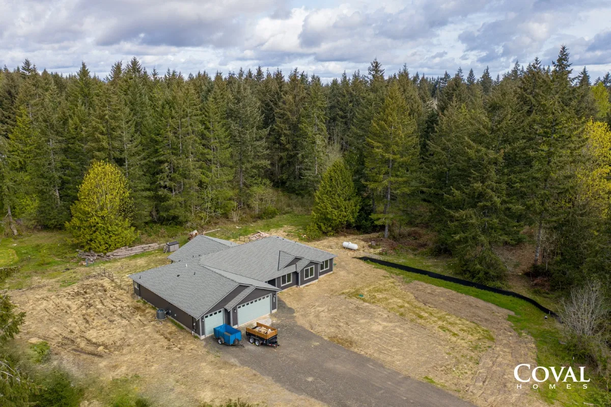 Aerial view of a gray-roofed house beside a forest, with a dirt driveway and blue bins.