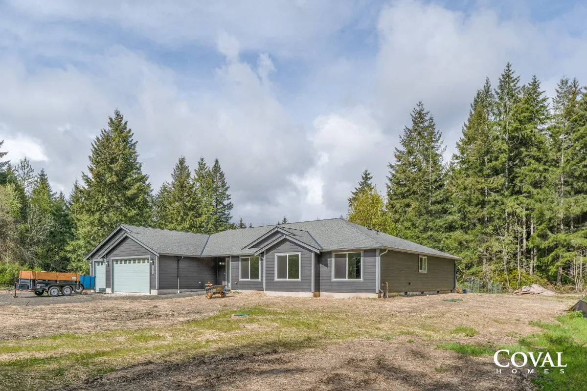 Modern gray house with attached garage, set in a clearing beside tall evergreen trees under a cloudy sky