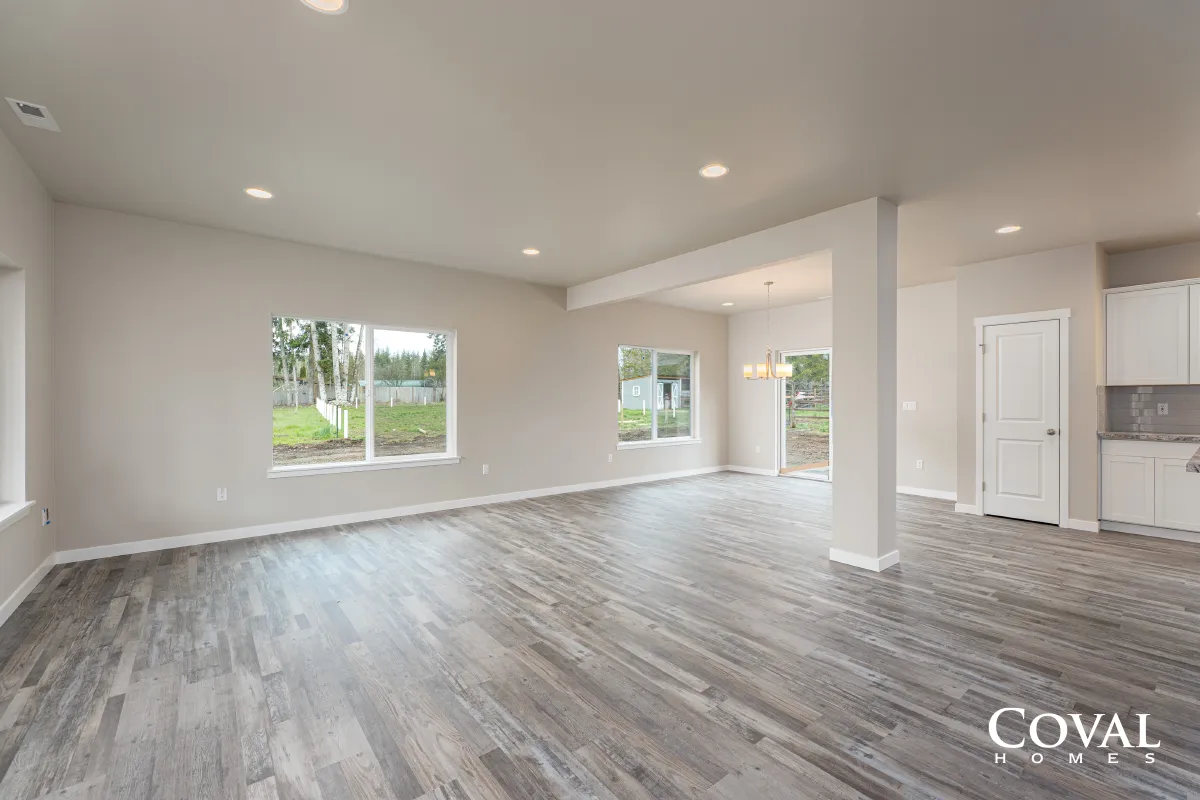Bright empty open-plan living room with gray wood floors, white walls, recessed lights, and windows overlooking greenery