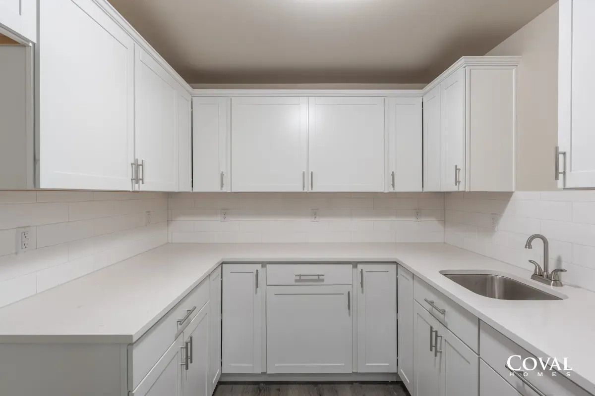 Bright white U-shaped kitchen with cabinets, countertops, and a stainless sink under a windowless wall.