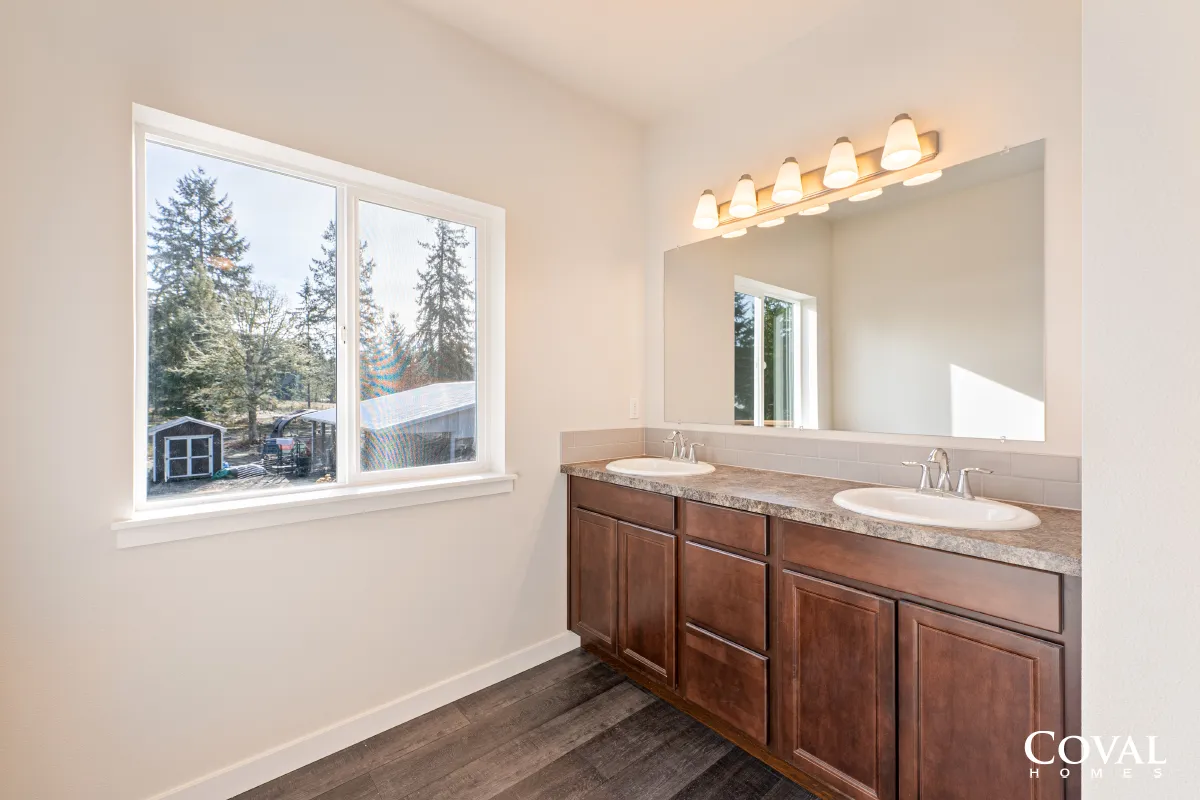 Bright bathroom with double vanity, large mirror, and window showing trees outside.