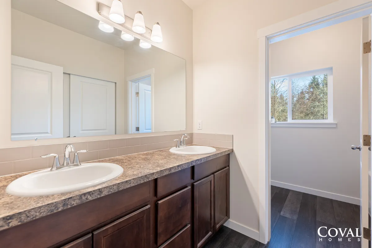 Bathroom vanity with double sinks, dark wood cabinets, mirror, and a window to a hallway or adjoining room