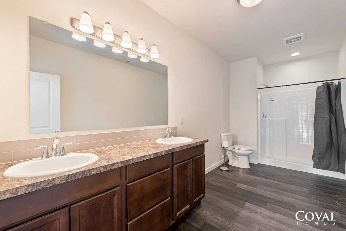 Bathroom with double vanity, large mirror, toilet, and shower curtain on wood-look flooring.