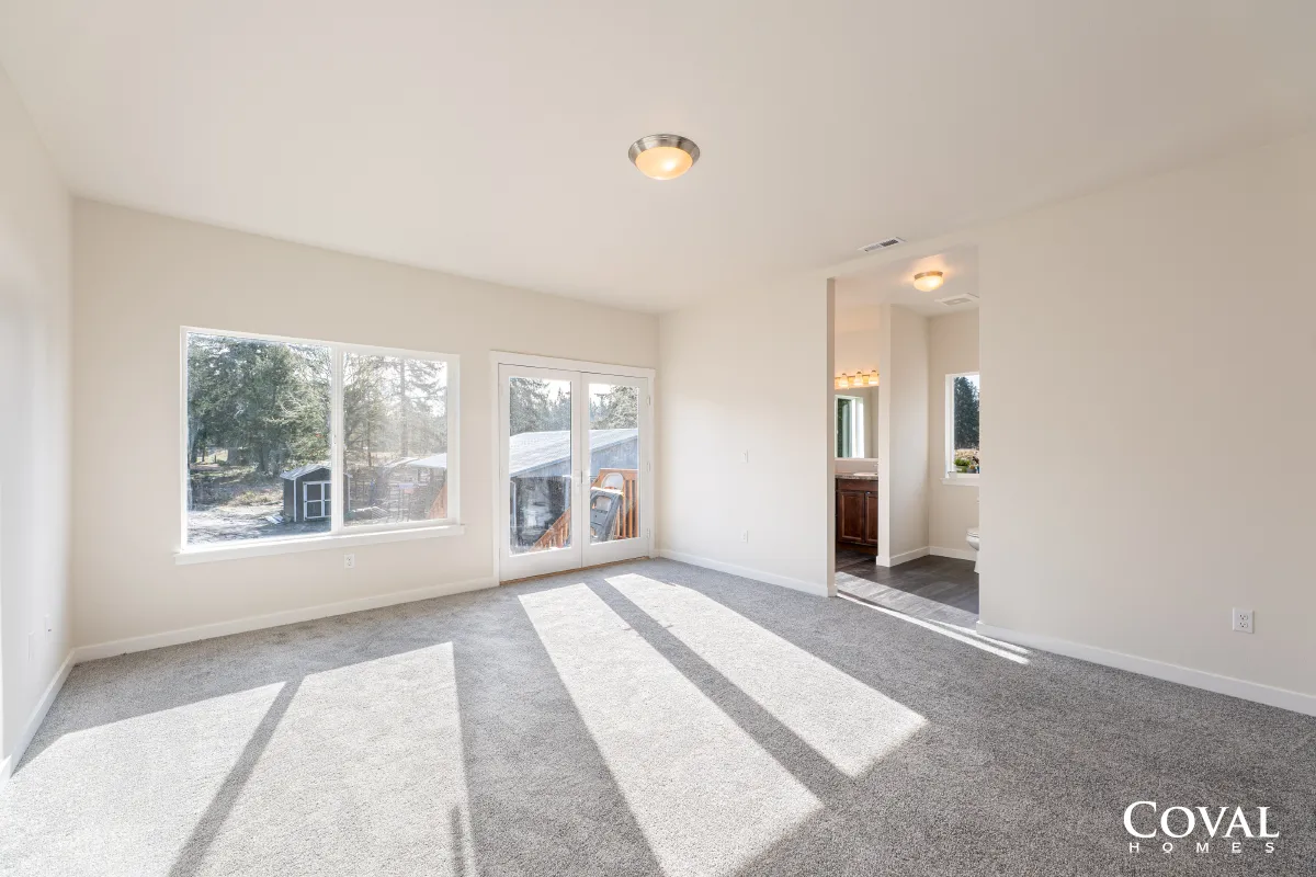 Bright empty living room with large windows, gray carpet, and sunlight casting long shadows
