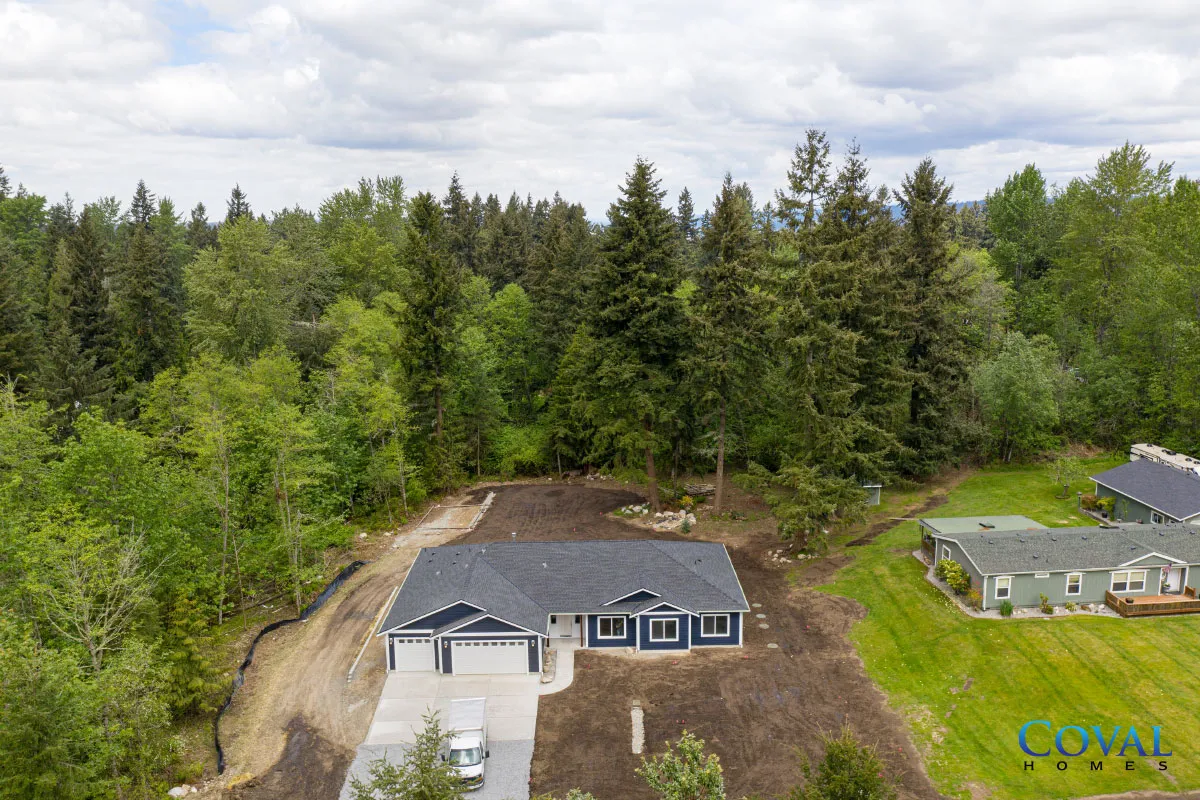 Aerial view of a house with a dark roof beside a dirt driveway, surrounded by dense green trees.