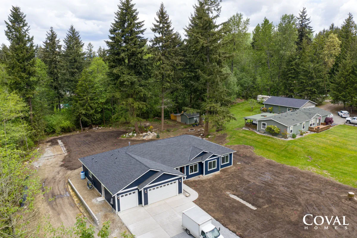 Aerial view of a new black-roofed house on a cleared lot beside a forest and neighboring homes.