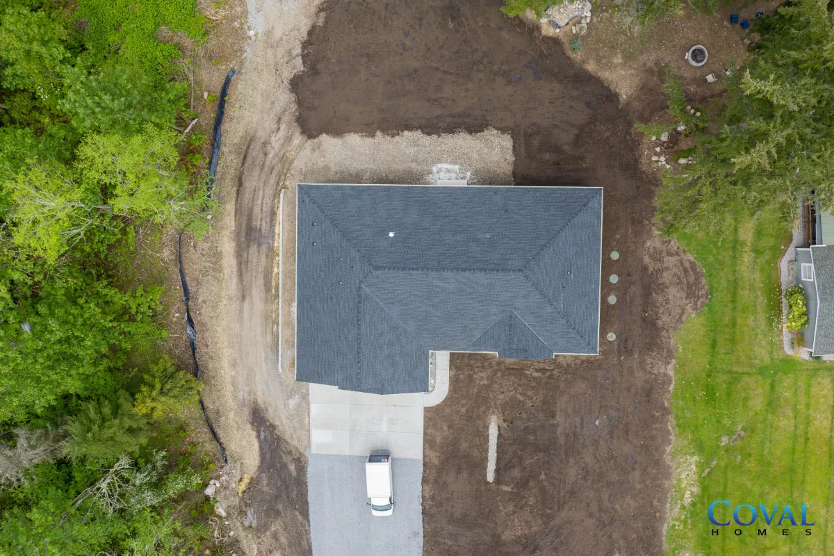 Aerial view of a house with a gray roof, driveway, and a parked white car beside a dirt yard.