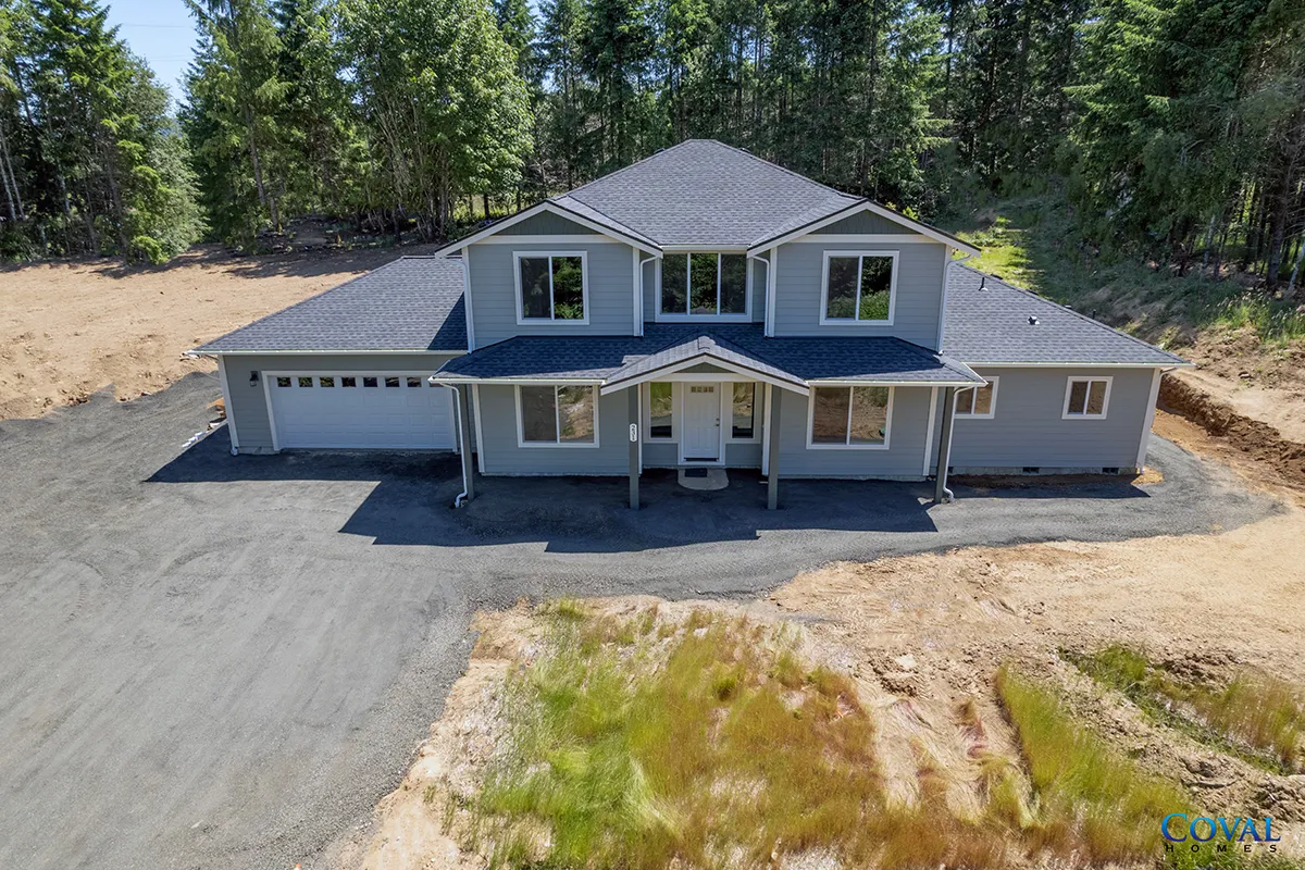 Large gray two-story house with a front porch and attached garage, surrounded by trees on a dirt lot