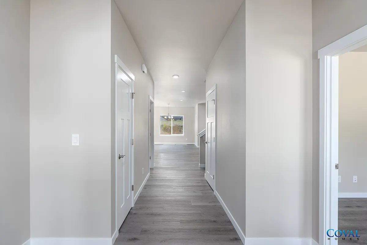 Bright hallway with gray wood floors, white walls, and doors leading to a sunlit room.