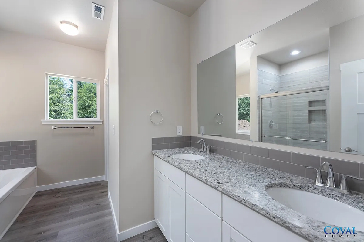 Bright bathroom with white double vanity, large mirror, gray tile floor, and window by a bathtub.