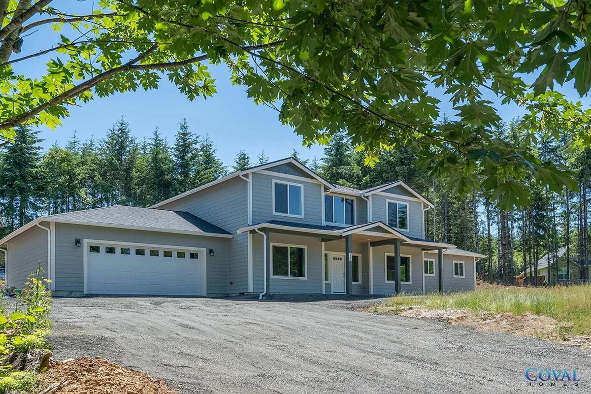 Two-story gray house with attached garage on a gravel lot, framed by green trees under a clear sky