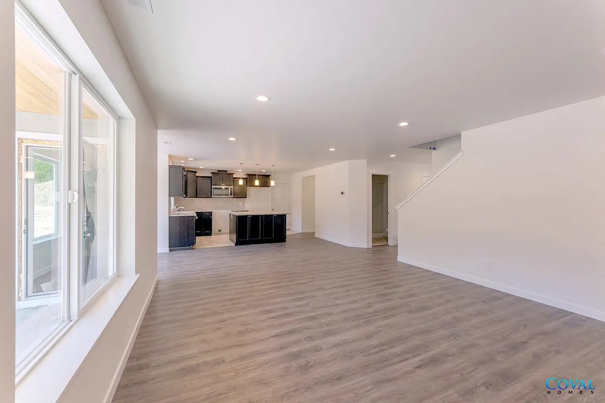 Empty modern living room with light wood floors, white walls, large windows, and a kitchen in the back