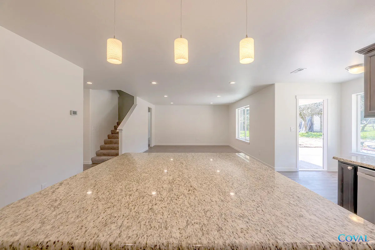 Spacious empty modern living room with granite island, pendant lights, staircase, and open doorway to outside