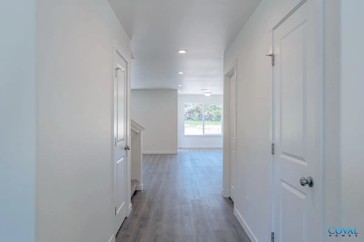 Bright modern hallway with white doors and trim, gray wood floors, and a large window at the end.