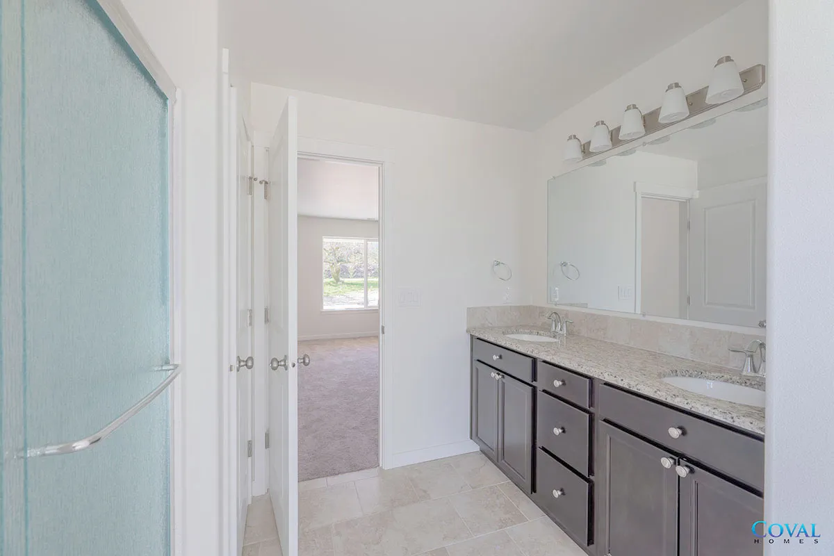 Bright bathroom with double vanity, large mirror, and light tile flooring, viewed toward an adjoining room.